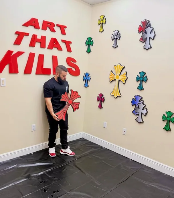 The artist behind ArtKr8tion in the studio holding a red epoxy cross, below the words ART THAT KILLS in red lettering, with a wall of coloured crosses beside him